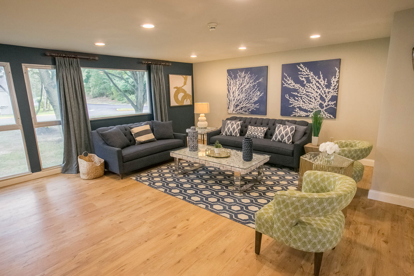 Light hardwood floor, a blue and white rug, blue couches, green and white chairs, a grey coffee table, a white wall, a blue wall with windows, and blue curtains