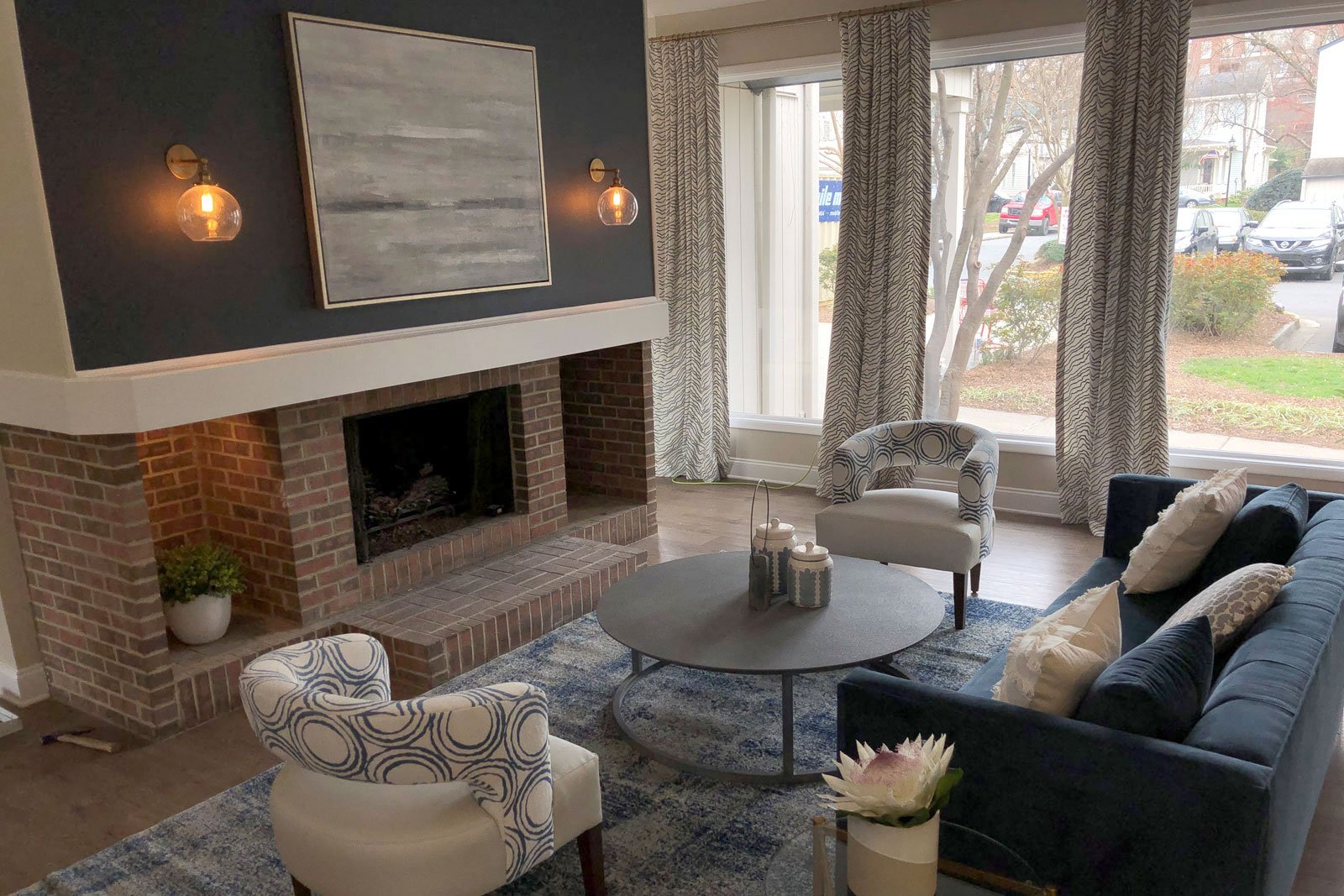 A wooden circular table sits in the middle of a patterned chair, velvet couch, brick fireplace and gold-rimmed painting with grey patterned cushions over the white windows
