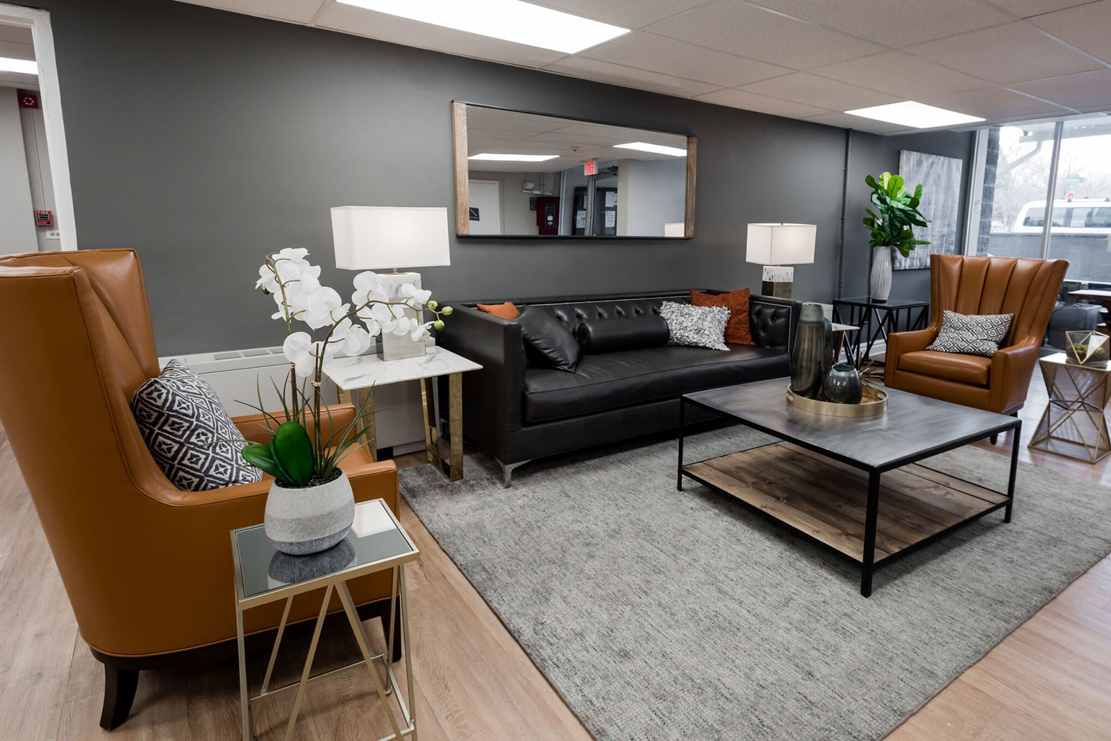 Wood flooring, a grey rug, a black couch, two brown chairs, a grey and black coffee table, dark grey walls, and a big mirror